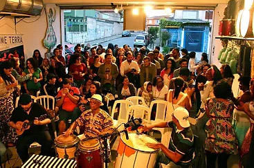 Grupo de pessoas reunido em um espaço cultural, músicos tocando instrumentos de percussão e cordas.