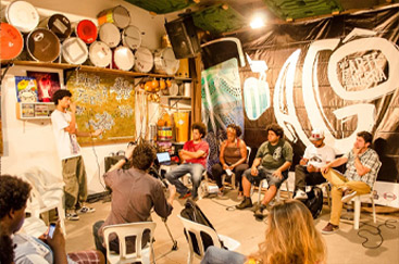 Roda de conversa em um espaço cultural com participantes sentados e tambores decorativos ao fundo.