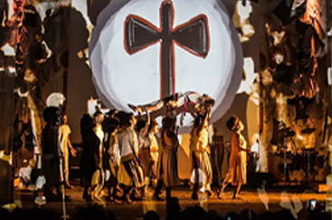 Apresentação teatral com grupo de atores em trajes tradicionais e fundo cruz estilizada.