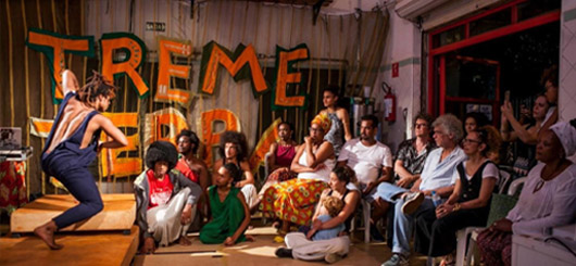 Apresentação artística em um espaço cultural, com uma performer no palco e um público diversificado assistindo.