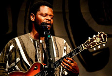 Músico tocando guitarra e cantando no palco, com expressão intensa e foco na performance.