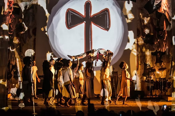 Apresentação teatral com grupo de atores em trajes tradicionais e fundo projetado com símbolo de uma cruz estilizada.