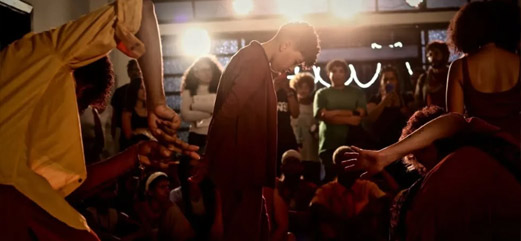 Grupo de performers em uma apresentação artística coletiva com público ao fundo em um espaço interno.