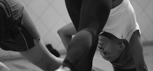 Dançarina em movimento acrobático, capturada em preto e branco, com expressão focada e pernas em destaque.