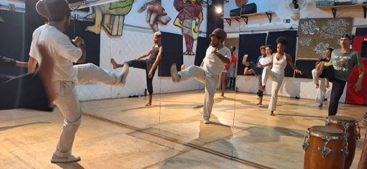 Grupo de dança praticando movimentos sincronizados em frente a espelhos em um estúdio com tambores e murais artísticos.