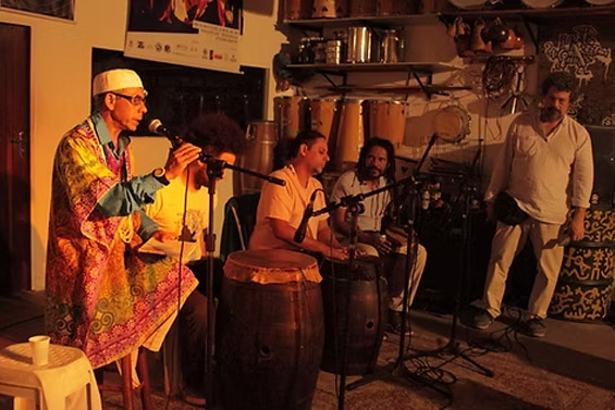 Grupo de músicos se apresentando em palco íntimo, com destaque para instrumentos de percussão e um cantor usando vestes coloridas.