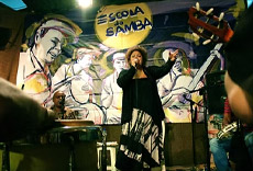 Cantora Fabiana Cozza se apresentando ao microfone, com músicos tocando ao fundo em um ambiente iluminado.