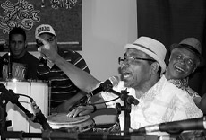 Mestre Kenura, com chapéu, tocando e se expressando ao microfone, enquanto outros músicos o acompanham no palco.