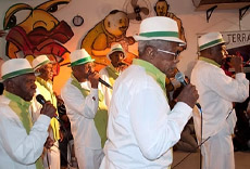 Grupo de músicos da velha guarda, com vestimentas brancas e chapéus, cantando ao microfone durante uma apresentação.