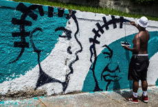 Artista de rua pintando um mural com imagens de rostos em um muro, utilizando tinta preta e azul.