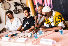 Mesa de debate com participantes discutindo em frente a instrumentos de percussão, com copos de água e microfone na mesa.
