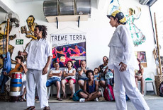 Dois atores se apresentam em uma peça de teatro enquanto o público, sentado ao fundo, observa atentamente.