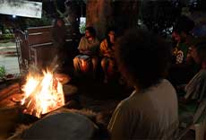 Grupo de pessoas reunidas em roda ao redor de uma fogueira, tocando instrumentos e conversando.
