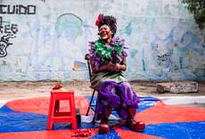 Artista palhaça sentada, sorrindo, em apresentação de rua sobre lona colorida.