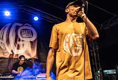 Rapper se apresentando no palco com microfone, enquanto DJ toca ao fundo.