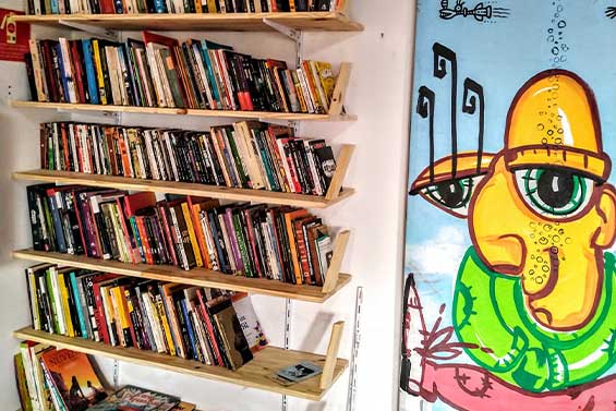 Estante de livros em espaço cultural, ao lado de um mural colorido com arte urbana.