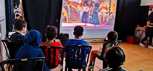 Grupo de crianças assistindo a um filme projetado em tela em espaço cultural.
