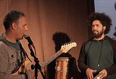 Dois músicos em apresentação, um tocando guitarra e outro segurando instrumento de percussão.