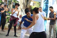 Duas pessoas jogando capoeira em espaço aberto, cercadas por público e músicos.