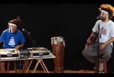 Dois músicos se apresentando em estúdio, um com toca-discos e outro com violão, ambos usando protetores faciais.